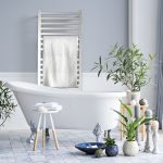 A modern bathroom features a white freestanding tub, green plants, and a rug. Towels rest on a small stool, while a towel hangs on the Quadro Q2142 16 Bar Hardwired Towel Warmer in Polished against a light gray wall bathed in natural light.