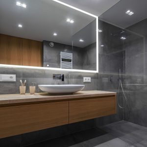 Modern bathroom with grey tiled walls, floating wood vanity, and walk-in glass shower. The Edge 84 Inch LED Lighted Bathroom Mirror hangs above a white vessel sink. Toothbrushes and a soap dispenser rest on the counter.