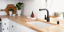 Modern farmhouse kitchen design with white cabinets, light wood countertops, black hardware, and a black faucet. Open shelf displays plants, jars, and decor. White tile backsplash enhances the minimal, clean look.