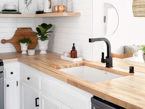 Modern farmhouse kitchen design with white cabinets, light wood countertops, black hardware, and a black faucet. Open shelf displays plants, jars, and decor. White tile backsplash enhances the minimal, clean look.