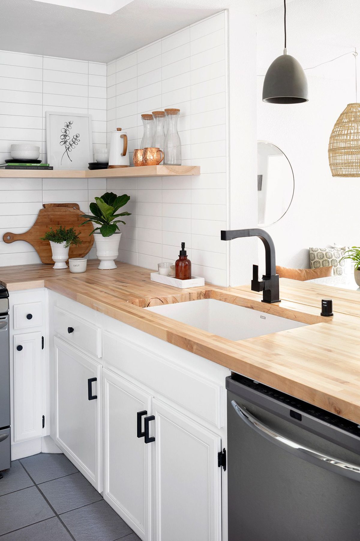 Modern farmhouse kitchen design with white cabinets, light wood countertops, black hardware, and a black faucet. Open shelf displays plants, jars, and decor. White tile backsplash enhances the minimal, clean look.
