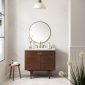 Modern bathroom with a round mirror above a wooden vanity with gold fixtures, marble countertop, and white walls. A small wooden stool with a towel and bottle sits to the left; decorative flowers are in the foreground.