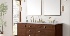 A modern bathroom with an Amberly 72" Double Vanity in Mid-Century Walnut with Eternal Jasmine Pearl top, two sinks, gold fixtures, rectangular mirrors, wall lights, a tall plant, and large windows for natural light.