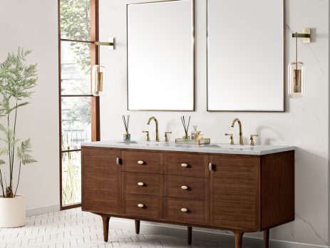 A modern bathroom with an Amberly 72" Double Vanity in Mid-Century Walnut with Eternal Jasmine Pearl top, two sinks, gold fixtures, rectangular mirrors, wall lights, a tall plant, and large windows for natural light.