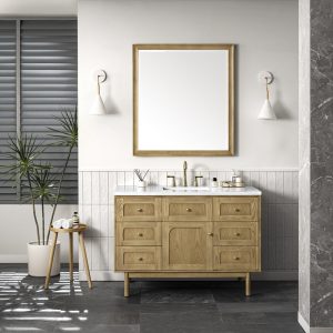 A modern bathroom features a Laurent 48" Single Bathroom Vanity Cabinet in Natural Oak, brass fixtures, a large framed mirror, white wall sconces, a potted plant, a towel-topped stool, and sleek gray tile flooring and walls.