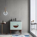 Modern bathroom with a sleek circular pendant light, VABURAG30FD 30-inch Aloe Green Freestanding Bathroom Vanity with a semicircular cutout, and a gold side table holding a soap dispenser and towel. A glass door reveals large containers outside, and a blue and white rug adorns the floor.