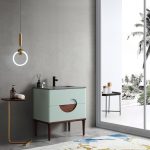 A modern bathroom features the Burano 30 inch Aloe Green Freestanding Bathroom Vanity with wooden legs, a round wall light, a small side table holding soap, and a large window displaying tropical palm trees outside.