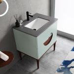 The Burano 30 inch Aloe Green Freestanding Bathroom Vanity features a light green cabinet with a circular cutout, gray countertop, rectangular white sink, black faucet, and is complemented by decorative items and a blue patterned rug.