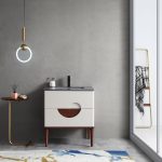 A minimalist bathroom showcases a gray wall and the Burano 30 inch Beige Freestanding Bathroom Vanity with circular wooden detail. A ring-shaped pendant light illuminates a small round table holding a book and brush. Nearby, an abstract-patterned rug enhances the floor beside a towel rack by the glass door.