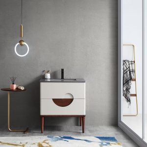 A minimalist bathroom showcases a gray wall and the Evano 30 inch Beige Freestanding Bathroom Vanity with circular wooden detail. A ring-shaped pendant light illuminates a small round table holding a book and brush. Nearby, an abstract-patterned rug enhances the floor beside a towel rack by the glass door.