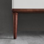 Close-up of an Burano 48 inch Beige Freestanding Bathroom Vanity leg, crafted from dark wood and attached to a white side panel, shown on a gray textured floor against a gray wall.