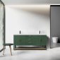 A modern bathroom showcases the Winchester 72 Forest Green Vanity with a white countertop and brass handles, flanked by two faucets. A round green stool with wooden legs is on the left by a large window, while a white toilet is secluded on the right behind a partial wall.