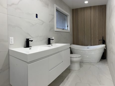 Modern bathroom with white marble walls and floor, featuring a Paterno 60" Double Wall Mount Vanity with black faucets, a toilet, a white freestanding bathtub, and a wooden slat accent wall behind the tub. A window sits above the tub.