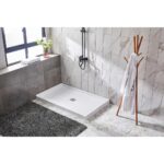 A modern bathroom features a Bhavana Single Threshold Shower Base, black shower fixture, marble wall tiles, a large window with a potted plant, a dark textured rug, and a wooden coat rack with a white robe.