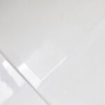 Close-up of the Bhavana Single Threshold Shower Base, white and textured, with a glossy transparent sheet partially covering it to highlight the contrast between matte and shiny surfaces.