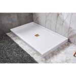 A Bhavana Single Threshold Shower Base with a gold square drain is installed in a bathroom with gray and white tiled walls and floors, next to a dark textured rug.
