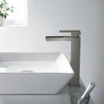 A modern bathroom sink features a rectangular white basin and the Quadra Waterfall Single Handle Bathroom Vessel Sink Faucet. In the background, a small plant sits on the windowsill, with a glass holding toothpaste on the counter.