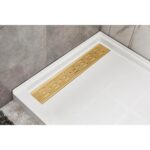 A modern shower floor featuring the Bhavana Single Threshold Shower Base with a rectangular white tray and gold linear drain cover, set against sleek gray and marble-patterned wall tiles.