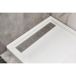 A close-up view of a modern shower floor with a white textured base and a rectangular stainless steel linear drain, surrounded by gray and marble-patterned tiles.