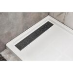 Close-up of a modern shower floor with the Bhavana Single Threshold Shower Base, featuring a rectangular stainless steel linear drain on light tiles and gray wall tiles. The white base displays a subtle grid pattern.