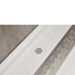 A close-up of the Bhavana Single Threshold Shower Base shows its white surface, central square drain, and a long rectangular silver grate on the right, with gray tiles on the adjacent wall.