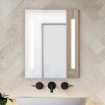 The Signature Royale 24"x36" Right Sided LED Lighted Mirror Medicine Cabinet features vertical LED lights, a digital clock in the lower right corner, and is paired with a minimalist black-faucet sink against beige-tiled walls.