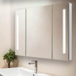 The Signature Royale 40 Inch x 36 Inch LED Lighted Mirror Medicine Cabinet features three mirrored doors, vertical LED light strips, a modern sink below, and a small green plant on the left side of the countertop.