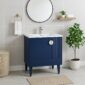 A modern bathroom featuring a Venezian 30 inch Bathroom Vanity With Sink, white countertop, round mirror, wall towel shelf, gold hand towel ring, potted plant, woven basket, and beige floor mat on light gray tiled walls and floor.
