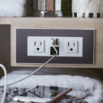 A smartphone charges on a bed via a cord plugged into a wall outlet panel with two sockets and two USB ports, next to stacked towels and bathroom items on a wooden shelf beneath a Linear 72 inch Single Bathroom Vanity Cabinet.
