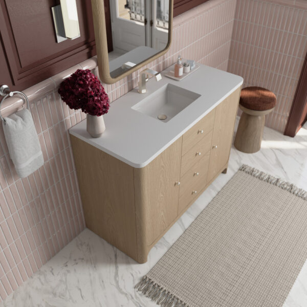 A bathroom with pink tiled walls and a marble floor showcases a Gracyn 48 inch Bathroom Vanity with white sink, complemented by a rectangular mirror, red flowers in a vase, and a brown stool. A beige fringed rug adorns the floor, while an ivory towel hangs on a silver holder.