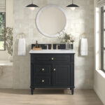 An elegant bathroom features the Lorelai 36 inch Bathroom Vanity Cabinet in Amber Birch, along with a white countertop and brass faucet. Above, a round mirror flanked by pendant lights hangs. Gold rings hold towels, while potted plants bring greenery to the cream tiled wall and hardwood floor.