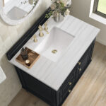 A bathroom vanity with a white marble countertop and a rectangular sink. Gold faucet fixtures, a wooden tray with a soap dispenser, and a vase with flowers decorate the surface. The vanity has dark cabinets and drawers, with a window nearby.