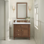 The Lorelai 36-inch Bathroom Vanity Cabinet features a wood finish and white countertop with gold accents. A rectangular mirror is paired with stylish wall sconces above, while a small bouquet decorates the counter and light hexagonal tiles enhance the floor.