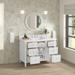 A Lorelai 48 inch Single Bathroom Vanity Cabinet with open drawers sits below a round mirror, flanked by wall sconces. Beige tiles surround the space, towels hang at the sides, and a nearby window lets in natural light.