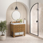The bathroom interior includes a Malibu 36 inch Bathroom Vanity Cabinet in Amber Birch, a round mirror, and potted plant. A black pendant light is above the sink, with towels inside the cabinet. Beige tiles outline the glass shower enclosure, and a vase of flowers sits on the counter.