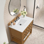 Bathroom vanity with a wooden cabinet, white countertop, and a black faucet. A round mirror hangs above, with a vase of white flowers beside soap and lotion dispensers. Tiled walls and a glass shower door are in the background.