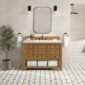 The bathroom features a Malibu 48 inch Bathroom Vanity Cabinet in Amber Birch, accented by a rectangular mirror and two black wall sconces overhead. White towels hang on either side, while a potted plant decorates the tiled floor near the freestanding bathtub.