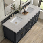 Modern bathroom vanity with a marble countertop, two square sinks, gold faucets, and black cabinets. Decorative plants and bathroom essentials are neatly arranged between the sinks. Wood floor and mirrors are visible.