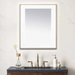 A bathroom vanity with a dark countertop, gold faucet, soap dispenser, and a single flower in a vase sits below the Lexington 30 inch x 36 inch LED Lighted Slim Frame Mirror on a light marble wall.