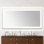 A modern bathroom features a Lexington 72 inch x 36 inch LED Lighted Slim Frame Mirror above a double-sink vanity with brass faucets, wooden cabinets, and minimal decor like a vase with white flowers and a reed diffuser.