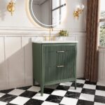A modern bathroom features the Aveline 30 Inch Freestanding Bathroom Vanity With Top in green with gold hardware, a white countertop, a round backlit mirror, gold wall sconces, black-and-white checkered floor tiles, and a small plant on the counter.