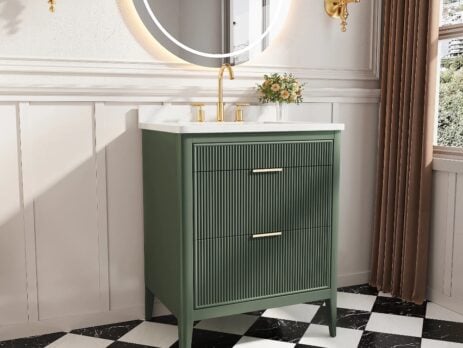 A modern bathroom features the Aveline 30 Inch Freestanding Bathroom Vanity With Top in green with gold hardware, a white countertop, a round backlit mirror, gold wall sconces, black-and-white checkered floor tiles, and a small plant on the counter.
