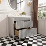 The Aveline 36 Inch Freestanding Bathroom Vanity With Top includes two open drawers, gold faucet, round mirror, small flower vase, wall sconce, and sits on a black-and-white checkered floor beside a window with brown curtains showing a city view.