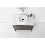Overhead view of the Celle 36 Inch Bathroom Vanity With Vessel Sink, showcasing a round white sink, minimalist faucet, two brown cabinet doors, a small bottle, and black cup on its white countertop beneath a partial circular mirror.