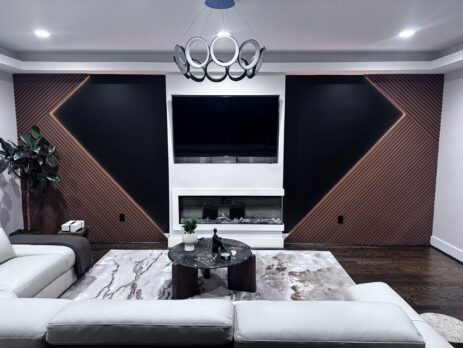 Modern living room with a white sectional sofa, round coffee table, wall-mounted TV above a fireplace, geometric wall panels, a plant, and a contemporary ring chandelier on the ceiling.