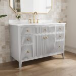 The Aveline 48 Inch Freestanding Bathroom Vanity With Top features a white marble countertop, gold faucet and handles, gray cabinets, a small flower vase under a gold-framed mirror, all set against a textured beige tile wall.