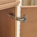 Close-up of a cabinet door with a silver metal hinge connecting it to the frame of the Linea 48 Inch Wall Mounted Bathroom Vanity, both made of light-colored wood.