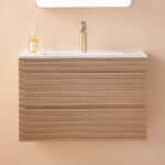 The Linea 30 Inch Wall Mounted Bathroom Vanity has a light wood slatted front, white countertop, gold faucet, and a reed diffuser bottle, all set against a beige wall.