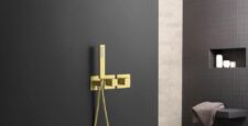 Modern bathroom with a matte black wall featuring a gold rain shower head, handheld shower, and faucet set. In the background, shelves hold toiletries and a towel rests on a wooden stool.