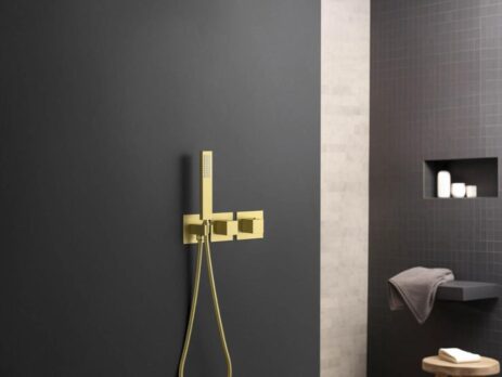 Modern bathroom with a matte black wall featuring a gold rain shower head, handheld shower, and faucet set. In the background, shelves hold toiletries and a towel rests on a wooden stool.